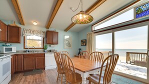 A kitchen with wooden cabinets, a dining table with chairs, a large window with a view of the beach, and a hanging light fixture.
