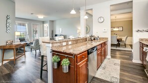 A modern kitchen with granite countertops, wooden cabinets, and a built-in dishwasher. There are potted plants on the island, and the room has hardwood flooring.