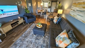 A modern living room with a sofa, ottoman, and coffee table, a TV on the wall, and a dining area in the background. - Condo # 102 Perfect 2 bedroom one bath condo directly across from  famous Siesta Key Beach. (Sarasota)