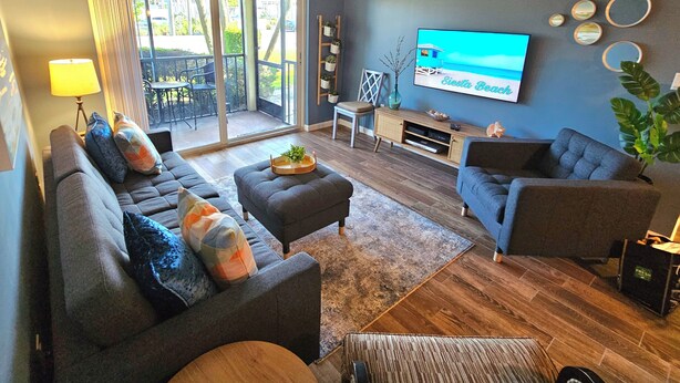 A living room with a sofa, ottoman, and armchair, a TV, and a sliding glass door leading to a balcony. - Condo # 102 Perfect 2 bedroom one bath condo directly across from  famous Siesta Key Beach. (Sarasota)