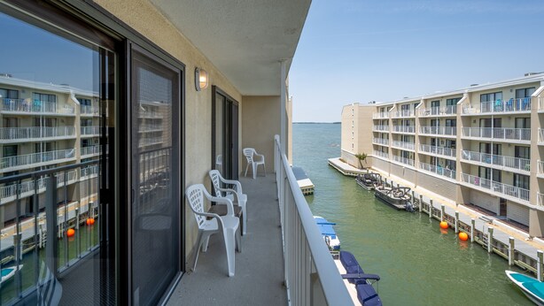 Een balkon met witte plastic stoelen, uitzicht op een jachthaven met boten, en een gebouw met balkons.