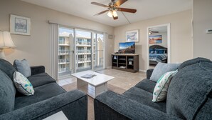 Een woonkamer met grijze banken, een witte salontafel en een plafondventilator. Er is een tv-meubel met een flatscreen televisie en een boekenplank. Een schuifdeur leidt naar een balkon met uitzicht op een gebouw.