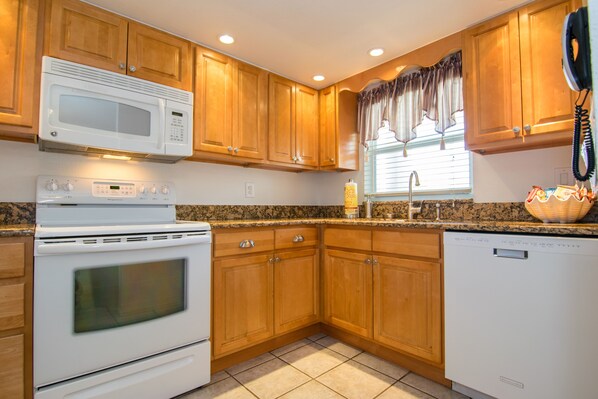 A kitchen with wooden cabinets, a microwave, stove, dishwasher, granite countertop, and a window with curtains. - Gulfsands 201 · Manatee Beach · Elevator · Pool (Holmes Beach)