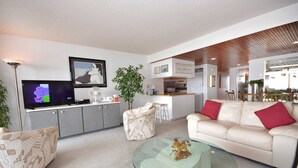 A modern living room with a sofa, a glass coffee table, a TV, and a kitchen area in the background.