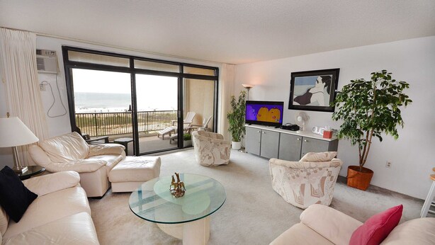 A living room with a glass coffee table, white sofas, a TV, and a large window overlooking the beach.