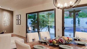 A dining area with a wooden table set for four, a chandelier, and a view of a pool through glass doors. - Andiamo al Sole - Palm Springs Luxury Retreat (Palm Springs)