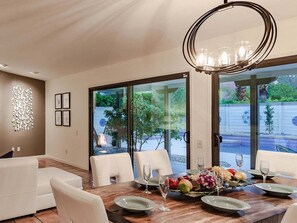 Un coin repas avec une table en bois dressée pour quatre personnes, un lustre et une vue sur une piscine, visible à travers des portes vitrées.
