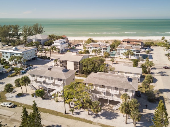 An aerial view of a coastal resort with multiple buildings, including a hotel with balconies and a beach in the background.