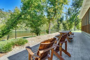 A wooden deck with chairs and tables overlooking a lake, surrounded by trees and a building with a wooden facade. - 'TroutFest' Riverfront Norfork Home, Great Fishing (Norfork)