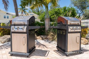 Deux barbecues d’extérieur sur roulettes, installés sur une surface pavée, avec des palmiers et une clôture en arrière-plan.