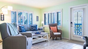 A living room with a grey sofa, a patterned armchair, and a coffee table. There are two windows with shutters, a door with a keypad lock, and a small table with a lamp.