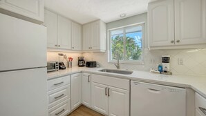 A kitchen with white cabinets, a dishwasher, a sink, and a window with a view of trees. - 4BR Coastal Cottage w/ Heated Private Pool | 1 Block to Bean Point Beach | Dog Friendly  *(See Note) ! (Anna Maria)