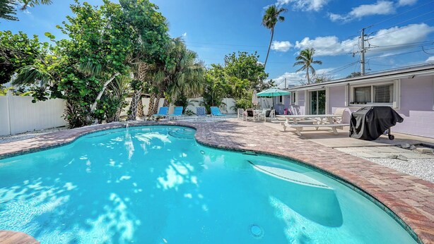 A swimming pool with a brick border, surrounded by lounge chairs and umbrellas, with a house and palm trees in the background. - 4BR Coastal Cottage w/ Heated Private Pool | 1 Block to Bean Point Beach | Dog Friendly *(See Note) ! (Anna Maria)