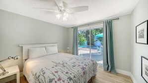 A bedroom with a bed, a ceiling fan, a bedside table with a lamp, and a view of an outdoor pool area. - 4BR Coastal Cottage w/ Heated Private Pool | 1 Block to Bean Point Beach | Dog Friendly  *(See Note) ! (Anna Maria)