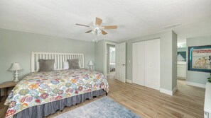 A bedroom with a bed, a ceiling fan, a nightstand, a lamp, a patterned bedspread, a gray skirted bed, a wooden floor, a gray rug, and a white door.