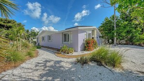 A single-story house with a light purple exterior, a gravel driveway, and a small garden with tropical plants. - 4BR Coastal Cottage w/ Heated Private Pool | 1 Block to Bean Point Beach | Dog Friendly  *(See Note) ! (Anna Maria)