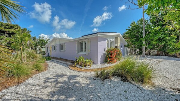 A single-story house with a light purple exterior, a gravel driveway, and a small garden with tropical plants.