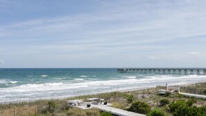 A coastal view with a pier extending into the ocean, a sandy beach, and a wooden boardwalk with seating.