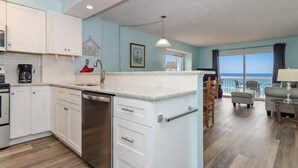 A modern kitchen with white cabinets, a dishwasher, and a countertop. There is a dining area with a table and chairs, and a view of the ocean through large windows. - First floor oceanfront condo in Ocean Dunes! (Kure Beach)