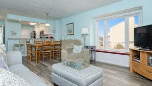 A modern living room with a sofa, ottoman, and a television. There is a dining area with a table and chairs, and a kitchen in the background. - First floor oceanfront condo in Ocean Dunes! (Kure Beach)