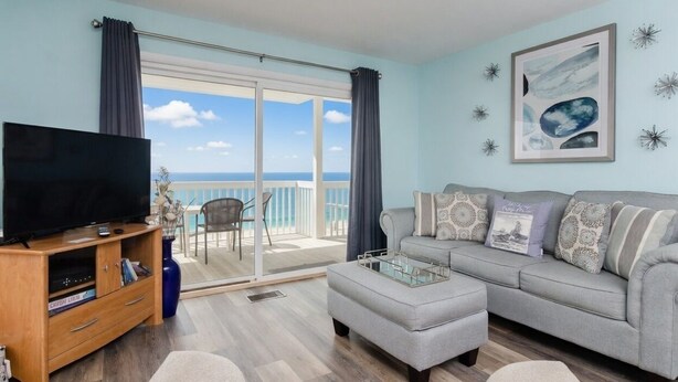 A living room with a television, a grey sofa, and a balcony with a view of the sea. - First floor oceanfront condo in Ocean Dunes! (Kure Beach)