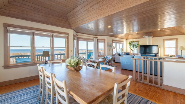 A spacious living area with a wooden ceiling, large windows offering a view of the beach, a dining table with chairs, and a kitchen area with a TV. - The Coast Is Clear - Corolla, Ocean Sands, Oceanfront, 150 Ft to Beach, Open All Year, Pets OK (Corolla)
