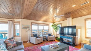 A spacious living room with a wooden ceiling, a flat-screen TV, comfortable seating, and large windows offering a view. - The Coast Is Clear - Corolla, Ocean Sands, Oceanfront, 150 Ft to Beach, Open All Year, Pets OK (Corolla)