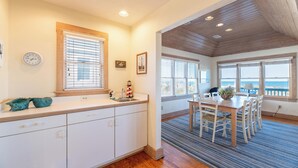 A kitchen with white cabinets, a wooden countertop, and a window with blinds. There is a dining area with a table and chairs, and a view of the ocean through large windows. - The Coast Is Clear - Corolla, Ocean Sands, Oceanfront, 150 Ft to Beach, Open All Year, Pets OK (Corolla)