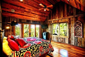 Un dormitorio de cabaña de madera con cama, ventilador de techo y ventanas grandes.