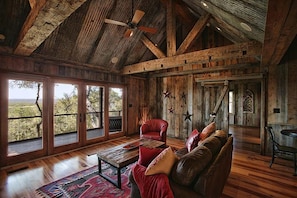 Un interior rústico con vigas de madera, un sofá de cuero y grandes ventanales con vistas al paisaje.