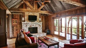 A rustic interior with a stone fireplace, wooden beams, a ceiling fan, and large windows. - Upscale Luxury Cabin! Has it all Hot tub, Fireplace, Gorgeous Views!!! (Wimberley)