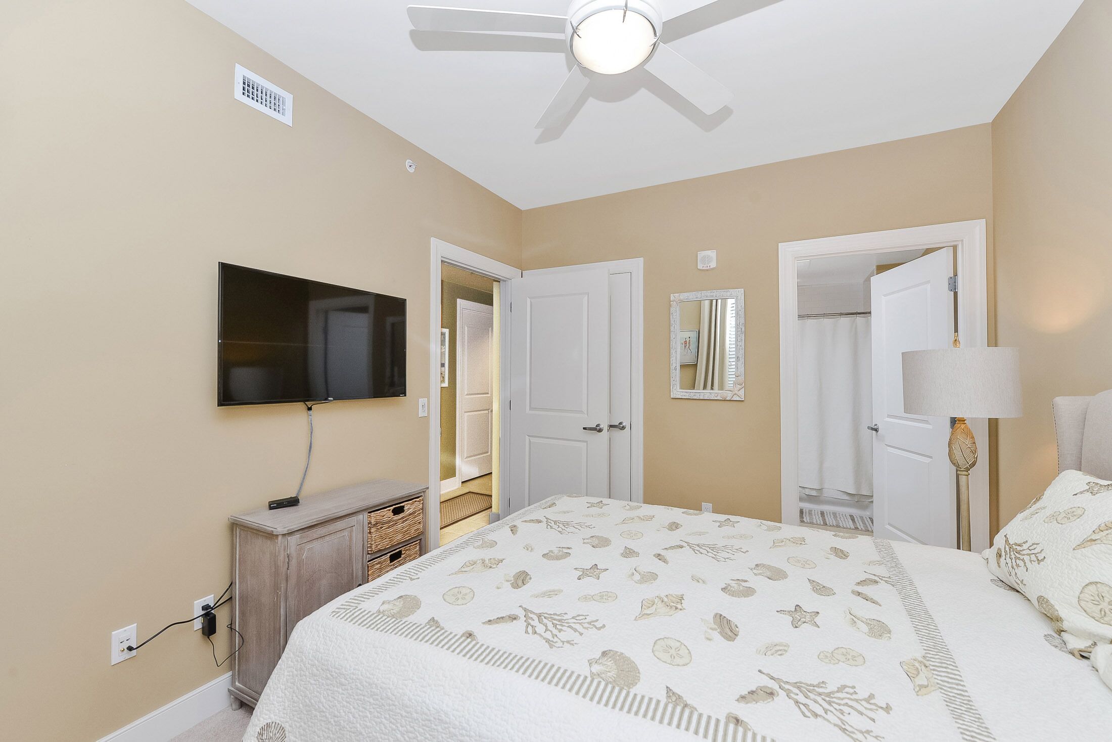 A bedroom with a bed, a television mounted on the wall, a ceiling fan, and two doors leading to other rooms.