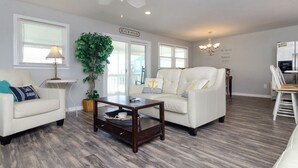 A living room with a white sofa, a coffee table, a ceiling fan, a potted plant, and a sign reading "BEACH HOUSE".