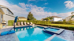 Une piscine dotée de chaises longues et d’un jacuzzi, le tout entouré d’une haie et de maisons.