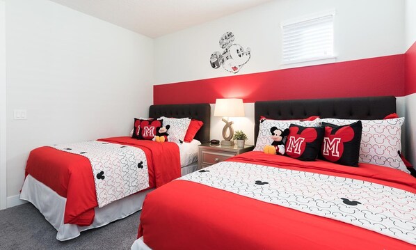 Ein Hotelzimmer mit zwei Betten, beide mit rotem und weißem Bettzeug mit Mickey Mouse Motiven. An einem Nachttisch steht eine Lampe und eine Mickey Mouse Figur.