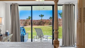 Habitación de hotel con cama, ventilador de techo, vista a un jardín y palmeras, y puerta corrediza de vidrio.