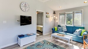 Ein Wohnzimmer mit blauem Sofa, weißem Couchtisch, Wand-Fernseher und großem Fenster mit Jalousien.