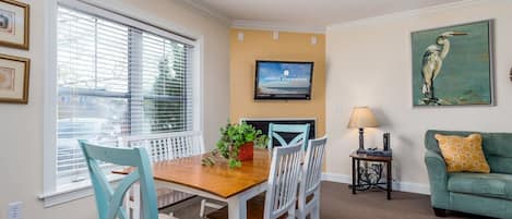 Un comedor con una mesa de madera y sillas blancas, un televisor en la pared y un sofá verde con un cojín amarillo.
