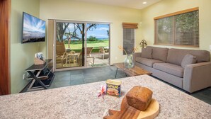 Ein Wohnzimmer mit Deckenventilator, Fernseher, einer Schiebetür zu einer Terrasse, einem Sofa und einer Küchenzeile mit einem Brot und einer Dose Spam.