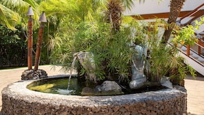 Ein runder Steinbrunnen mit Wasser, umgeben von tropischen Pflanzen und Palmen.