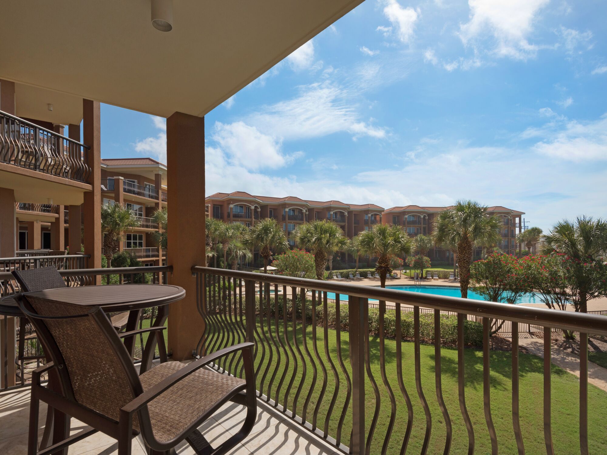 A balcony with a view of a swimming pool, palm trees, and a building complex.