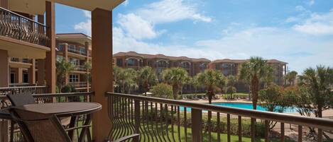 A balcony with a view of a swimming pool, palm trees, and a building complex.