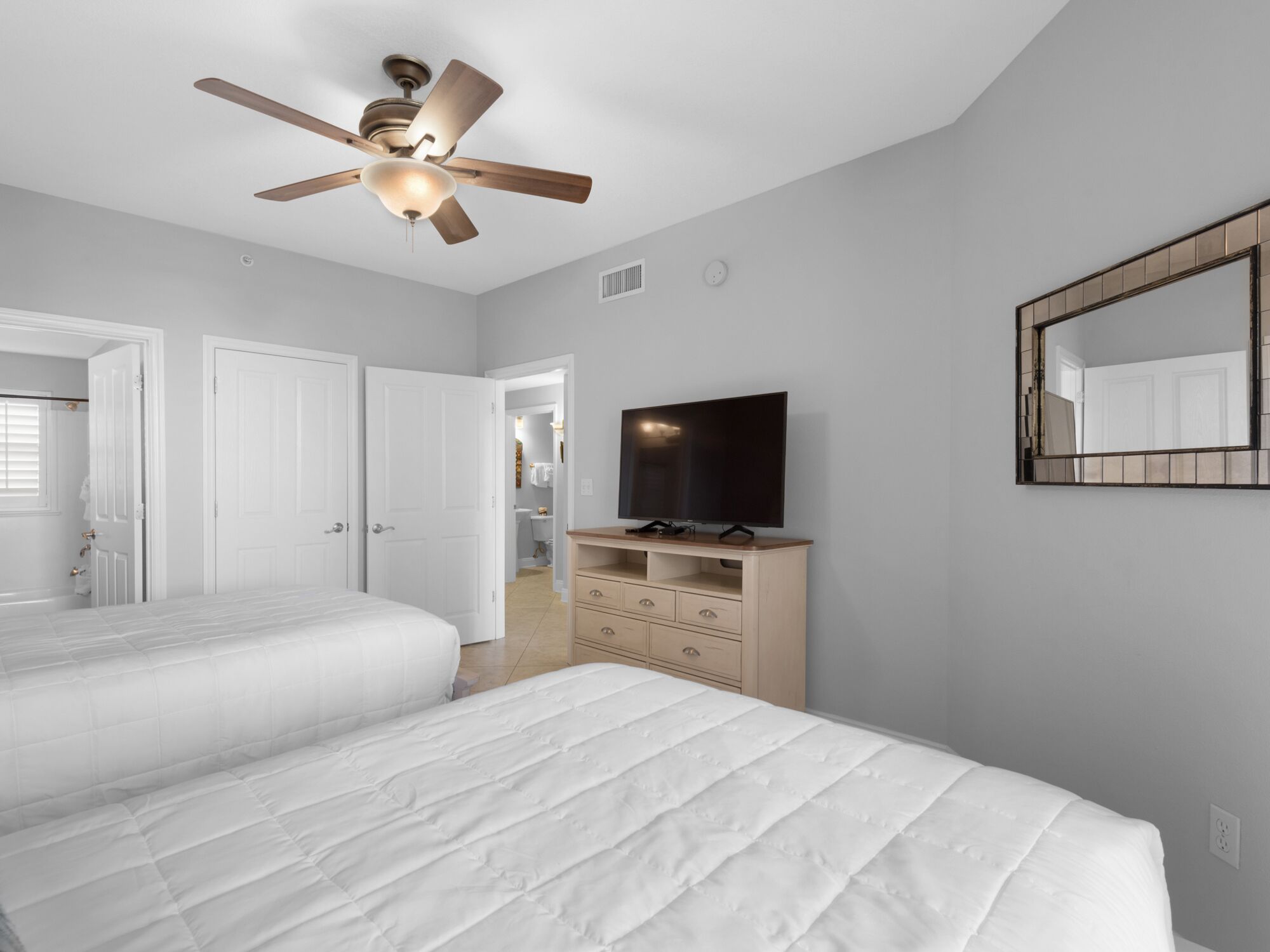 A bedroom with two beds, a ceiling fan, a TV on a wooden dresser, and a large mirror on the wall.