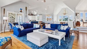 A spacious living room with a blue sofa set, a white ottoman, and a coffee table. There is a ceiling fan, a large window with a view, and a kitchen area in the background. - V1233- Beautiful! 5 BRs, Private Pool, Hot Tub, Pool Table and Dog Friendly! (Corolla)