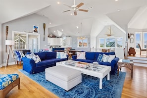 A spacious living room with a blue sofa set, a white ottoman, and a coffee table. There is a ceiling fan, a large window with a view, and a kitchen area in the background. - V1233- Beautiful! 5 BRs, Private Pool, Hot Tub, Pool Table and Dog Friendly! (Corolla)