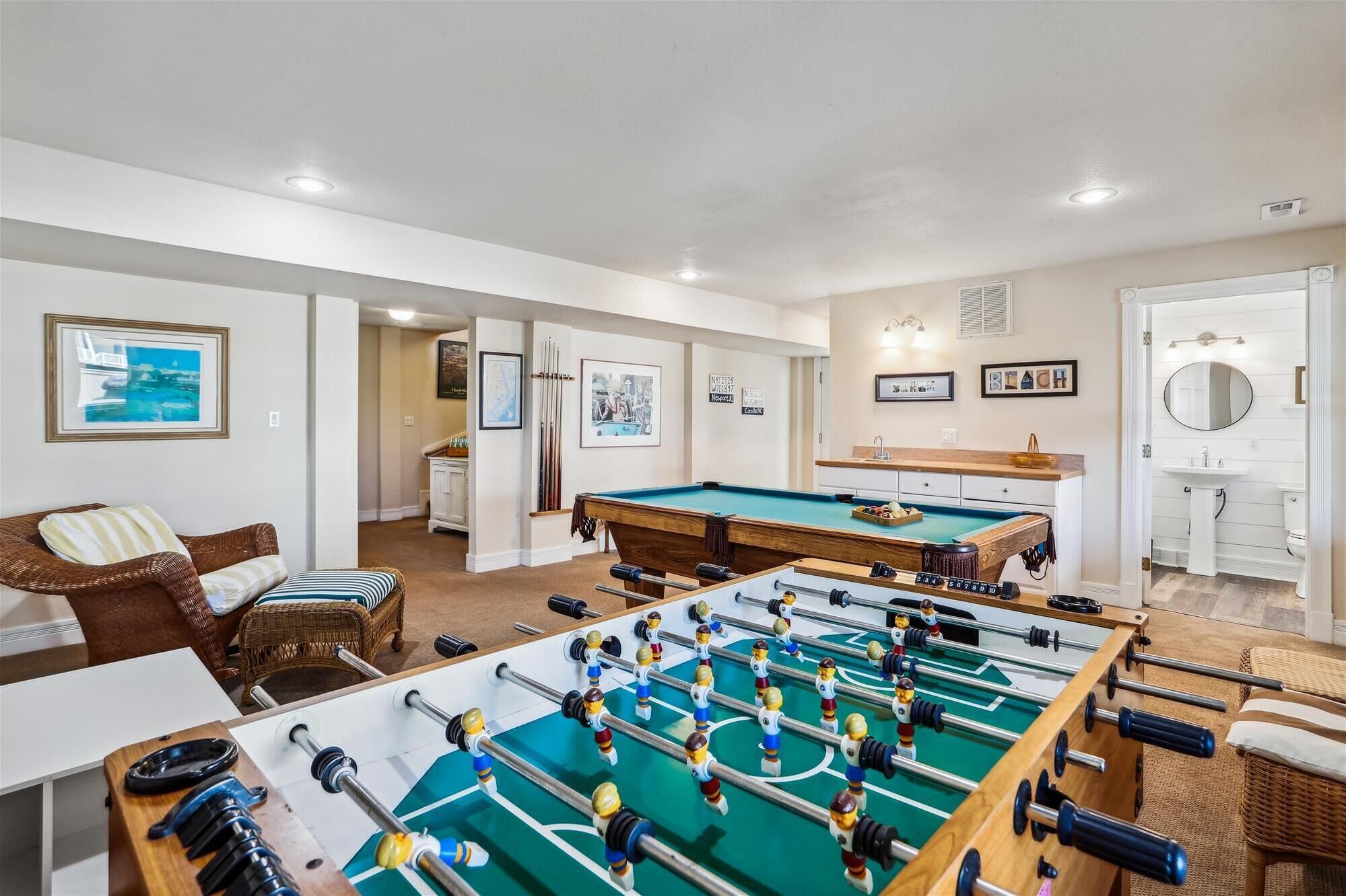 A room with two foosball tables, a billiards table, a sofa, and a bathroom with a mirror.