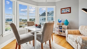 A corner room with a dining table and chairs, a wicker side table with decorative items, a framed picture on the wall, and large windows offering a view of houses and the beach. - V1233- Beautiful! 5 BRs, Private Pool, Hot Tub, Pool Table and Dog Friendly! (Corolla)