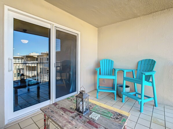 A balcony with a sliding glass door, two turquoise Adirondack chairs, a small wooden table, and a lantern.