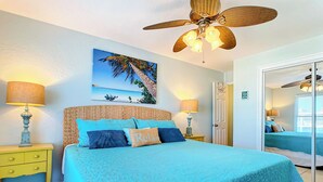 A bedroom with a bed, two bedside tables, a ceiling fan, a painting of a palm tree, and a door leading to another room. - Surfs up in this Cute 2bed/2ba condo! Ocean view! No drive Beach in NSB (New Smyrna Beach)