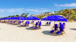 A beach with rows of blue beach chairs and umbrellas, a clear sky, and a building in the background. - 312 Breakers: Sleeps 4, Free Bikes, Pool, Oceanfront (Hilton Head Island)